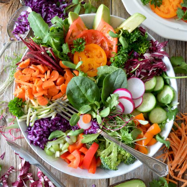 Eine bunte Salatschüssel gefüllt mit Spinat, Tomaten, Avocado, Karotten, Radieschen, Gurken, Rotkohl und Paprika. Um die Schüssel herum stehen Salatzutaten, Utensilien, Olivenöl und Avocado auf einem rustikalen Holztisch.