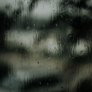 Ein Fenster, das mit Regentropfen und Schlieren bedeckt ist, mit einer unscharfen Ansicht von Bäumen und der freien Natur im Hintergrund, die eine stimmungsvolle, atmosphärische Szene schafft.