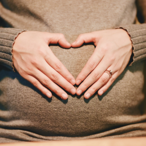 Eine Person, die einen Pullover trägt, legt ihre Hände auf den schwangeren Bauch und formt mit den Fingern eine Herzform.