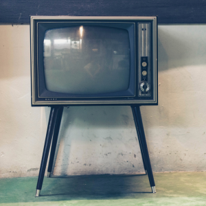 Ein alter Fernseher mit Drehknöpfen und vier Beinen steht vor einer hellen, strukturierten Wand auf einem grünlichen Boden. Der Bildschirm des Fernsehers ist leer und reflektiert etwas Licht.