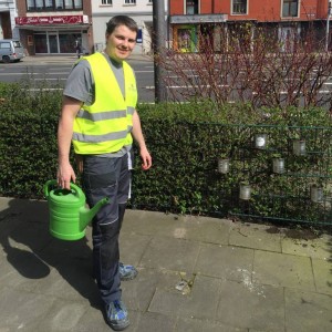 Ein Mann mit einer gelben Sicherheitsweste steht an einem sonnigen Tag im Freien und hält eine grüne Gießkanne in der Nähe von Sträuchern und Kerzen, mit Gebäuden und einer Straße im Hintergrund.