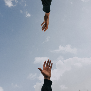 Zwei sich entgegenstreckende Hände vor einem blauen Himmel mit vereinzelten Wolken; die untere Hand streckt sich nach oben, während die obere Hand nach unten reicht, wobei in der rechten unteren Ecke Bäume zu sehen sind.