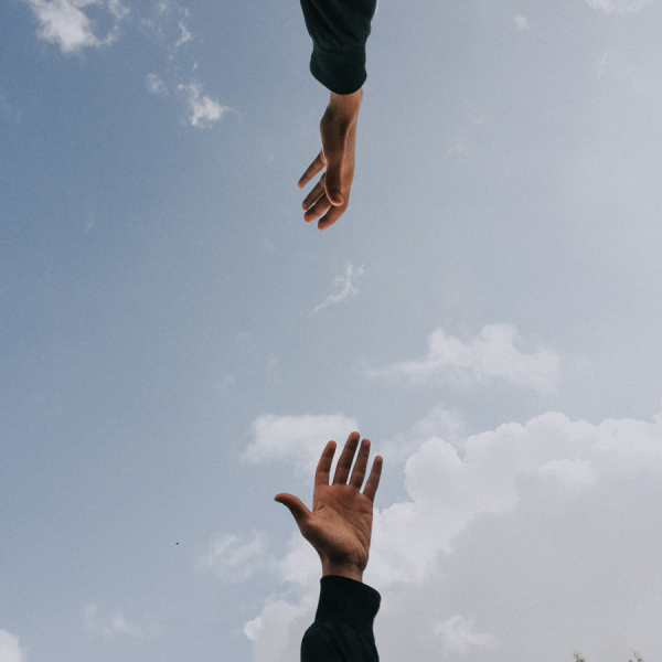 Zwei sich entgegenstreckende Hände vor einem blauen Himmel mit vereinzelten Wolken; die untere Hand streckt sich nach oben, während die obere Hand nach unten reicht, wobei in der rechten unteren Ecke Bäume zu sehen sind.