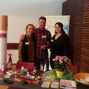 In einem Raum mit holzgetäfelten Wänden stehen drei Personen hinter einem Tisch, auf dem Haushaltsprodukte, Broschüren und eine Topfpflanze liegen. Sie lächeln und tragen Namensschilder, was auf eine Gemeinschafts- oder Informationsveranstaltung schließen lässt.