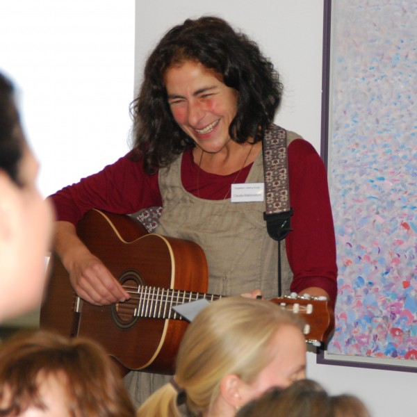Eine Frau mit lockigem Haar lächelt, während sie vor einer Gruppe eine akustische Gitarre spielt. Eine andere Frau steht daneben und lächelt ebenfalls. Hinter ihnen hängt ein buntes abstraktes Gemälde an der Wand.
