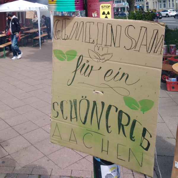 Auf einem handgeschriebenen Schild an einem Laternenpfahl steht "GEMEINSAM für ein SCHÖNEREB AACHEN". Bunte Blumentöpfe und Luftballons schmücken den Pfosten. Im Hintergrund versammeln sich Menschen bei Bänken und einem roten Gebäude unter einem weißen Zelt.