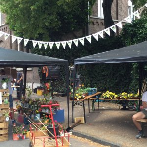 Straßenmarktszene mit zwei überdachten Zelten, in denen Blumen und Pflanzen ausgestellt sind. Menschen sitzen an Tischen, und zur Dekoration gehören ein Wimpel und Holzpaletten. Im Hintergrund sind ein Backsteingebäude und eine mit Efeu bewachsene Mauer zu sehen.