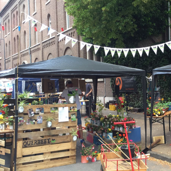 Straßenmarktszene mit zwei überdachten Zelten, in denen Blumen und Pflanzen ausgestellt sind. Menschen sitzen an Tischen, und zur Dekoration gehören ein Wimpel und Holzpaletten. Im Hintergrund sind ein Backsteingebäude und eine mit Efeu bewachsene Mauer zu sehen.