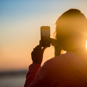 Eine Person im Kapuzenpulli macht ein Foto mit einem Smartphone bei Sonnenuntergang, wobei die Sonne hell scheint und einen warmen, leuchtenden Silhouetteneffekt erzeugt.