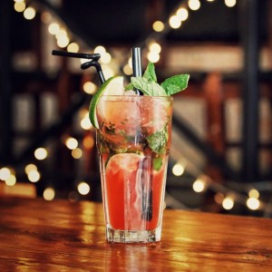 Ein Glas mit einem roten Cocktail mit Minzblättern, Limettenscheiben und einem Strohhalm steht auf einem Holztisch, während im verschwommenen Hintergrund dekorative Lichterketten leuchten.