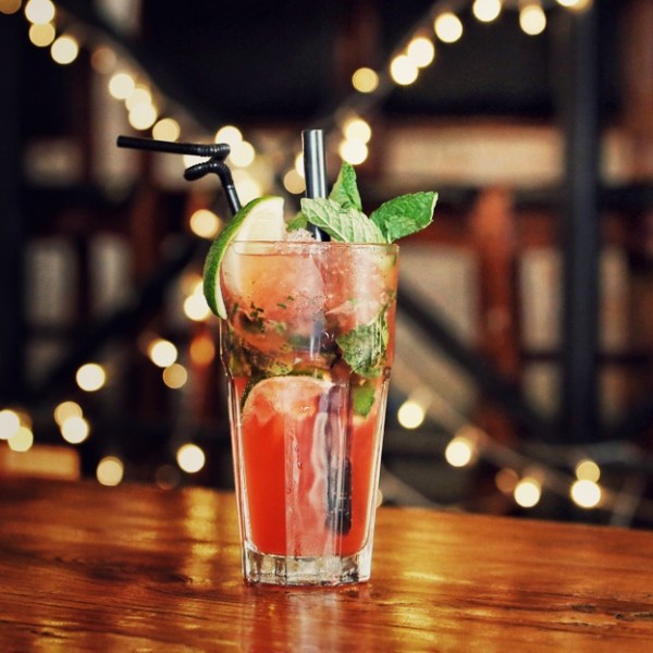 Ein Glas mit einem roten Cocktail mit Minzblättern, Limettenscheiben und einem Strohhalm steht auf einem Holztisch, während im verschwommenen Hintergrund dekorative Lichterketten leuchten.
