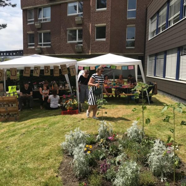 Menschen versammeln sich unter weißen Zelten bei einer Gemeinschaftsveranstaltung im Freien neben den Gebäuden. Im Vordergrund befindet sich ein Garten mit Blumen, und an einem Zelt hängt ein QUERBEET-Banner. Einige Menschen sitzen, während eine Person in der Nähe steht.