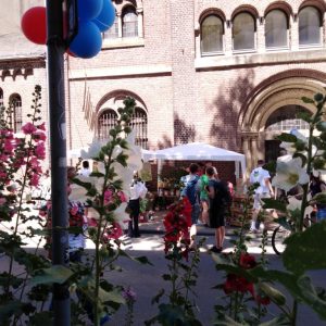 Menschen versammeln sich in der Nähe eines Backsteingebäudes mit gewölbten Fenstern, das durch hohe, blühende Blumen hindurch zu sehen ist. Ein weißes Zelt ist aufgebaut, und an einer Stange sind rote und blaue Luftballons befestigt, was auf eine Veranstaltung oder Feier im Freien hindeutet.