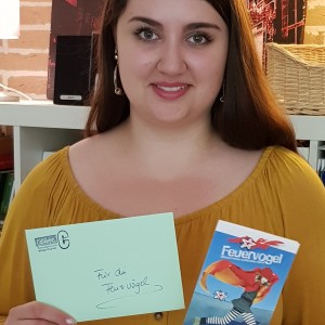 Eine Frau mit langen braunen Haaren und senfgelber Bluse hält einen grünen Umschlag und ein "Feuervogel"-Büchlein in der Hand. Sie steht in einem Haus mit Regalen und Büchern im Hintergrund.