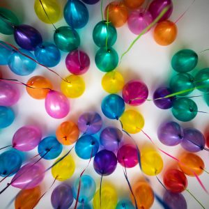 Bunte Luftballons in Blau-, Violett-, Rosa-, Gelb-, Grün- und Orangetönen sind von oben betrachtet in einem kreisförmigen Muster vor einem weißen Hintergrund angeordnet.