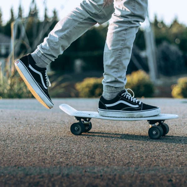Nahaufnahme einer Person in schwarzen und weißen Turnschuhen und hellen Hosen, die auf einem weißen Skateboard auf einer gepflasterten Fläche im Freien fährt, mit Grün und unscharfem Hintergrund.