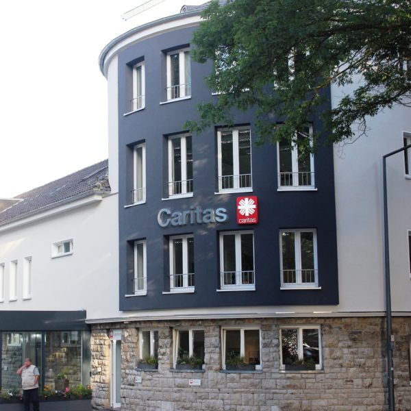 Ein modernes Gebäude mit dunklen und hellen Wänden, die das Logo und den Namen der Caritas tragen. Eine Person steht in der Nähe des Eingangs, und große Fenster säumen die oberen Stockwerke. Davor stehen ein Baum und eine Straßenlaterne.