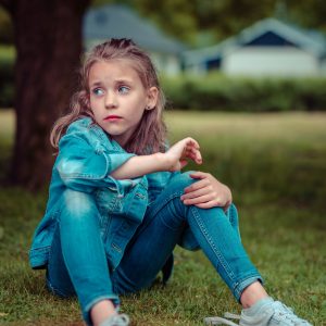 Ein junges Mädchen in Jeansjacke und Jeans sitzt im Gras und schaut mit nachdenklichem Blick zur Seite. Sie befindet sich im Freien, mit verschwommenen Bäumen und Gebäuden im Hintergrund.