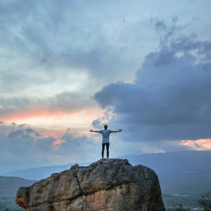 Eine Person steht mit ausgestreckten Armen auf einem großen Felsen und blickt auf einen dramatischen Himmel bei Sonnenauf- oder -untergang, umgeben von fernen Bergen und verstreuten Wolken.