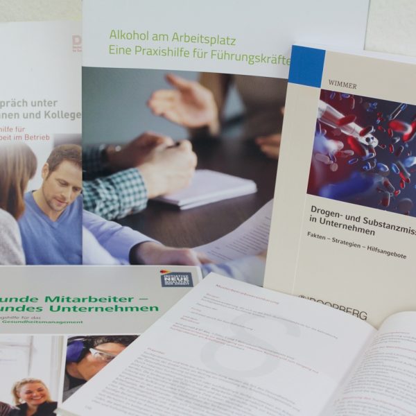 Auf einem Tisch liegt eine Sammlung von deutschsprachigen Broschüren und Büchern zum Thema Gesundheit am Arbeitsplatz aus, die sich mit Themen wie Alkohol, Drogenmissbrauch, Mitarbeitergesundheit und Kommunikation zwischen Kollegen befassen.