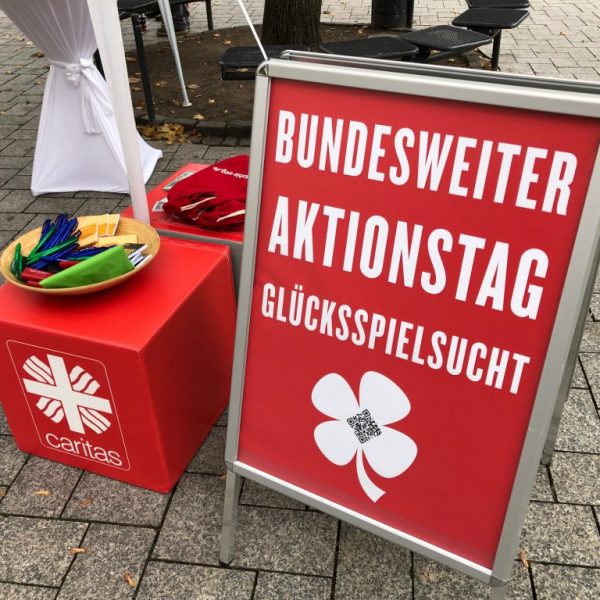Ein rotes Schild an einer Straße mit der Aufschrift Bundesweiter Aktionstag Glücksspielsucht steht neben einer Caritas-Box mit einer Schale mit bunten Stiften und Flyern. Im Hintergrund sind Stühle und ein weißes Zelt zu sehen.
