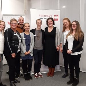 Acht Menschen stehen lächelnd vor einem Transparent mit der Aufschrift "Suchthilfe Aachen". Sie befinden sich in einem gut beleuchteten Raum, gekleidet in legerer Business-Kleidung. Die Gruppe wirkt fröhlich und hilfsbereit.