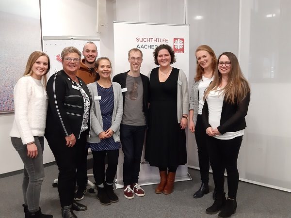 Acht Menschen stehen lächelnd vor einem Transparent mit der Aufschrift "Suchthilfe Aachen". Sie befinden sich in einem gut beleuchteten Raum, gekleidet in legerer Business-Kleidung. Die Gruppe wirkt fröhlich und hilfsbereit.