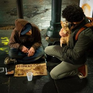 Eine Person sitzt auf einem nassen Bürgersteig mit einem HOMELESS-Schild, einer Tasse und etwas Essen. Eine andere Person kniet in der Nähe, hält einen kleinen Hund und sieht die Person auf dem Boden an. Es ist Nacht und die Straße ist feucht.