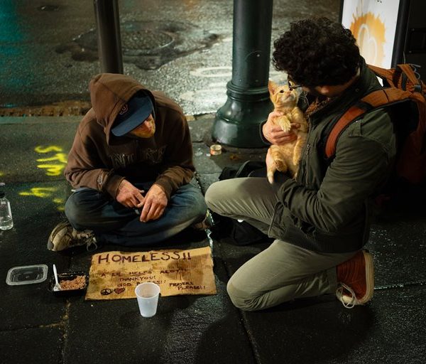 Eine Person sitzt auf einem nassen Bürgersteig mit einem HOMELESS-Schild, einer Tasse und etwas Essen. Eine andere Person kniet in der Nähe, hält einen kleinen Hund und sieht die Person auf dem Boden an. Es ist Nacht und die Straße ist feucht.
