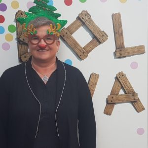 Eine lächelnde Person trägt eine festliche Brille mit Rentiergeweih, eine rote Nase und eine Weihnachtsbaummütze und steht vor einer gepunkteten Wand mit dem Schriftzug HOLLA in Holzbuchstaben.