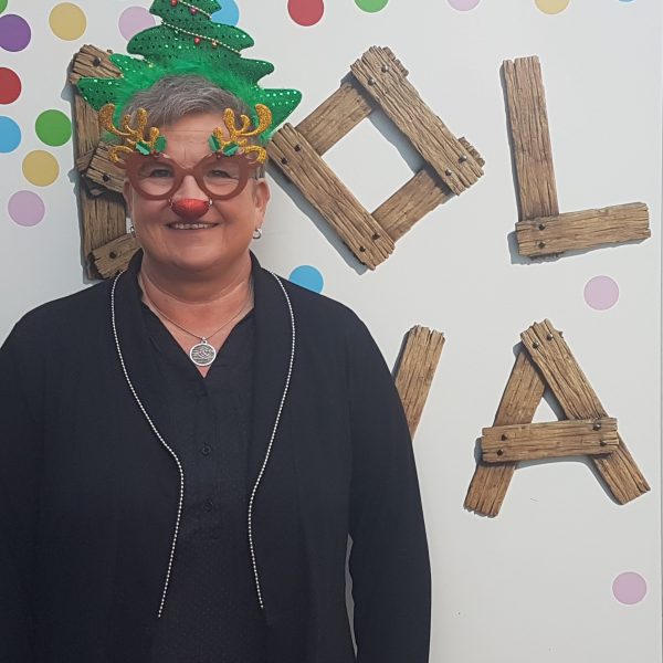 Eine lächelnde Person trägt eine festliche Brille mit Rentiergeweih, eine rote Nase und eine Weihnachtsbaummütze und steht vor einer gepunkteten Wand mit dem Schriftzug HOLLA in Holzbuchstaben.