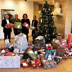 Vier Personen stehen vor einem geschmückten Weihnachtsbaum mit einem großen Stapel eingepackter Geschenke vor ihnen. Im Innenbereich sind eine Büste und gerahmte Kunstwerke an der Wand hinter ihnen zu sehen.
