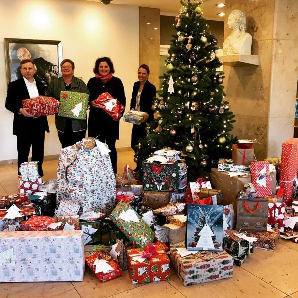 Vier Personen stehen vor einem geschmückten Weihnachtsbaum mit einem großen Stapel eingepackter Geschenke vor ihnen. Im Innenbereich sind eine Büste und gerahmte Kunstwerke an der Wand hinter ihnen zu sehen.