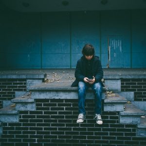 Ein Junge in dunkler Jacke und Jeans sitzt allein auf einer Backsteintreppe im Freien und schaut auf sein Handy. Um ihn herum liegen abgefallene Blätter, und im Hintergrund ist ein schummriges, leeres Gebäude zu sehen.