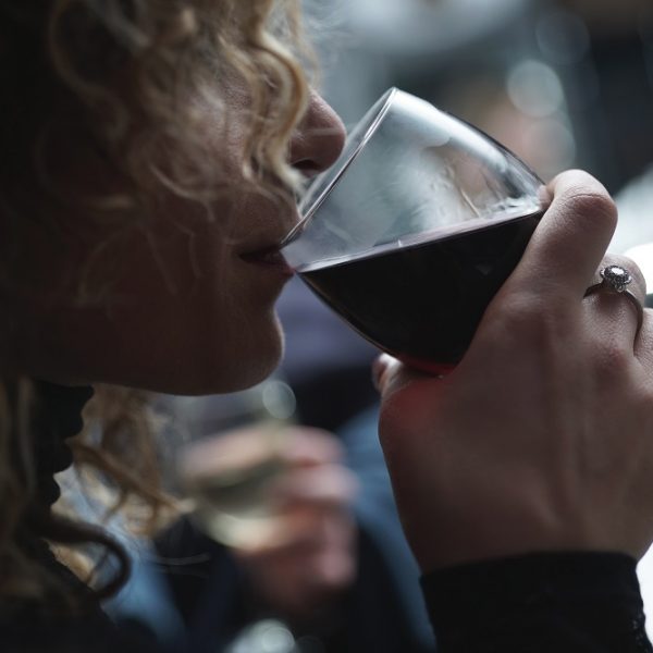 Eine Person mit lockigem Haar hält ein Glas Rotwein in der Hand und nippt daran. Sie trägt einen Ring am Finger; der Hintergrund ist leicht unscharf und suggeriert eine entspannte oder gesellige Atmosphäre.