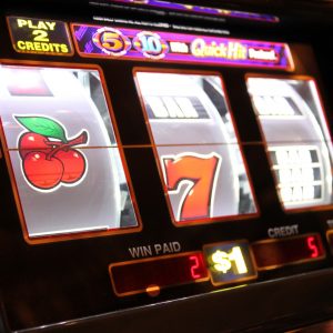 Nahaufnahme eines Spielautomaten mit sich drehenden Walzen, die eine Kirsche, eine rote Sieben und ein unscharfes drittes Symbol zeigen. Die Tasten des Automaten, die Guthabenanzeige und die Anzeige für den Einsatz von 1 $ sind sichtbar.