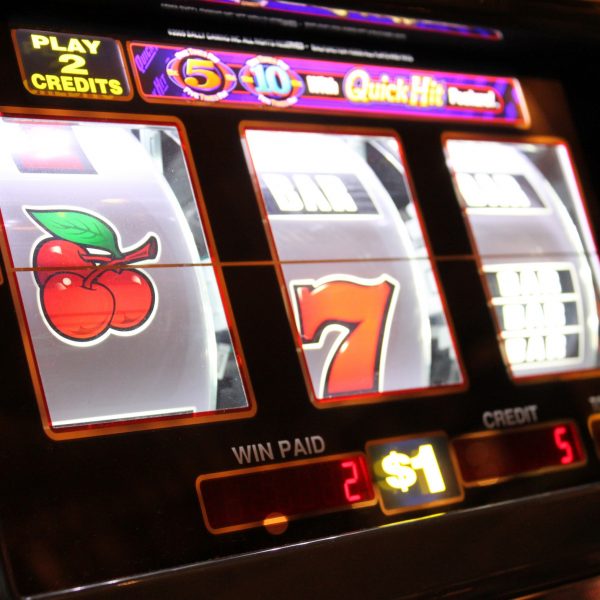Nahaufnahme eines Spielautomaten mit sich drehenden Walzen, die eine Kirsche, eine rote Sieben und ein unscharfes drittes Symbol zeigen. Die Tasten des Automaten, die Guthabenanzeige und die Anzeige für den Einsatz von 1 $ sind sichtbar.