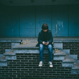 Ein Junge sitzt allein auf einer Betontreppe und blickt auf das Telefon in seinen Händen. Er trägt eine dunkle Jacke, Jeans und Turnschuhe. Der Hintergrund ist blau und einfarbig, mit einigen gefallenen Blättern auf den Stufen.