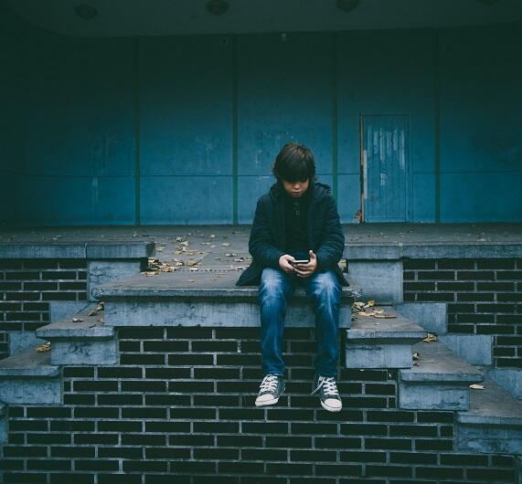 Ein Junge sitzt allein auf einer Betontreppe und blickt auf das Telefon in seinen Händen. Er trägt eine dunkle Jacke, Jeans und Turnschuhe. Der Hintergrund ist blau und einfarbig, mit einigen gefallenen Blättern auf den Stufen.