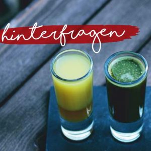 Zwei Schnapsgläser auf einer dunklen Fläche, eines mit gelber und das andere mit dunkelgrüner Flüssigkeit gefüllt, auf einem Holztisch. Darüber steht in weißer Schrift auf einem roten Pinselstrich das deutsche Wort "hinterfragen".