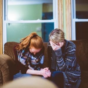 Zwei Frauen sitzen eng beieinander auf einer Couch, den Kopf gesenkt und halten sich an den Händen, um sich zu trösten. Die eine bedeckt ihr Gesicht mit der Hand, was darauf hindeutet, dass sie einen Moment der Traurigkeit oder emotionalen Unterstützung teilen.