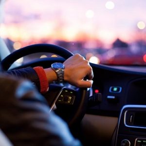 Eine Person, die eine Uhr trägt, fährt ein Auto in der Dämmerung, wobei die Lichter der Stadt vor der Windschutzscheibe verschwimmen. Im Vordergrund sind das Armaturenbrett und das Lenkrad zu sehen.