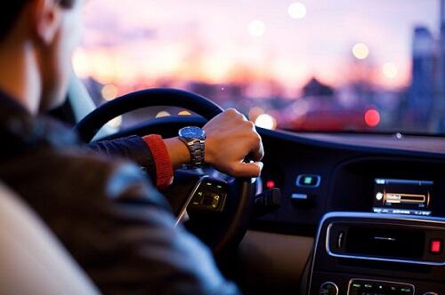 Eine Person, die eine Uhr trägt, fährt ein Auto in der Dämmerung, wobei die Lichter der Stadt vor der Windschutzscheibe verschwimmen. Im Vordergrund sind das Armaturenbrett und das Lenkrad zu sehen.