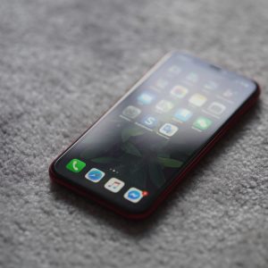 Ein Smartphone mit roter Hülle liegt mit der Vorderseite nach oben auf einem strukturierten grauen Teppich, auf dem der Startbildschirm mit verschiedenen App-Symbolen zu sehen ist.