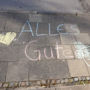 Die Kreidezeichnung auf einem Bürgersteig lautet "ALLES Gute" in Blau und Rosa, mit einem gelben Herz auf der linken und einem grünen vierblättrigen Kleeblatt auf der rechten Seite.