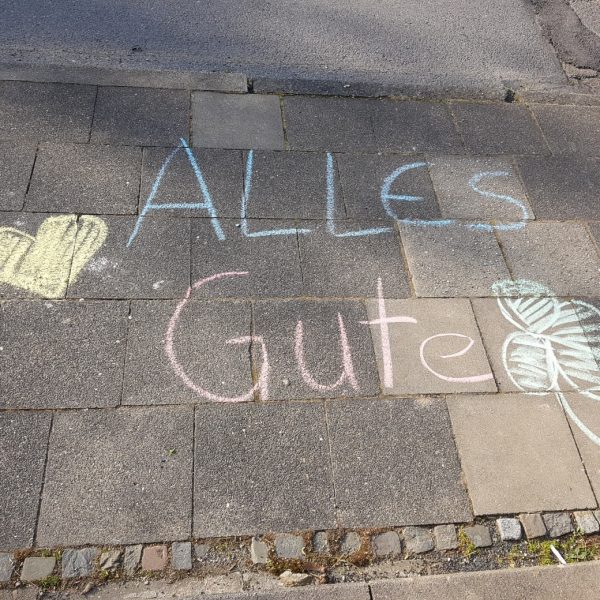 Die Kreidezeichnung auf einem Bürgersteig lautet "ALLES Gute" in Blau und Rosa, mit einem gelben Herz auf der linken und einem grünen vierblättrigen Kleeblatt auf der rechten Seite.