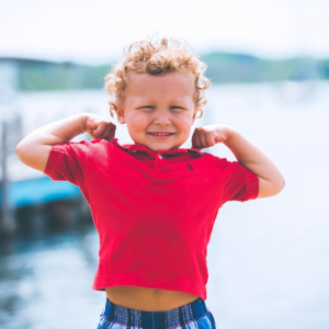 Ein kleiner Junge mit lockigem Haar und rotem Hemd lächelt und bewegt spielerisch seine Arme am Wasser, mit verschwommenen Booten und Bäumen im Hintergrund.