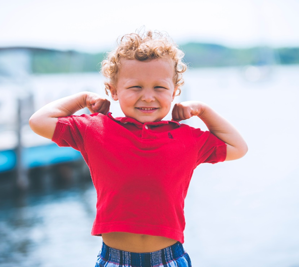 Ein kleiner Junge mit lockigem Haar und rotem Hemd lächelt und bewegt spielerisch seine Arme am Wasser, mit verschwommenen Booten und Bäumen im Hintergrund.