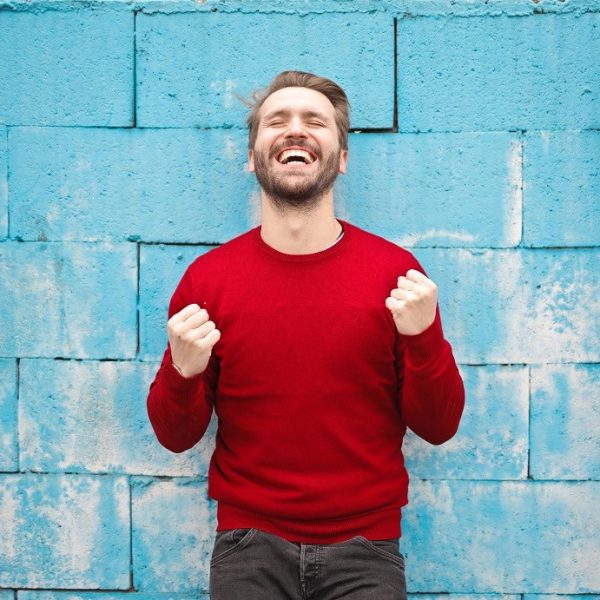 Ein Mann in einem roten Pullover steht vor einer blauen Backsteinmauer, lächelt breit, hat die Augen geschlossen und die Fäuste vor Aufregung oder Freude geballt.