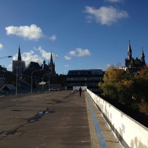 Eine Person geht allein auf einem weiten, leeren Dach oder Parkplatz unter einem blauen Himmel mit vereinzelten Wolken, mit gotischen Kirchtürmen und städtischen Gebäuden im Hintergrund.